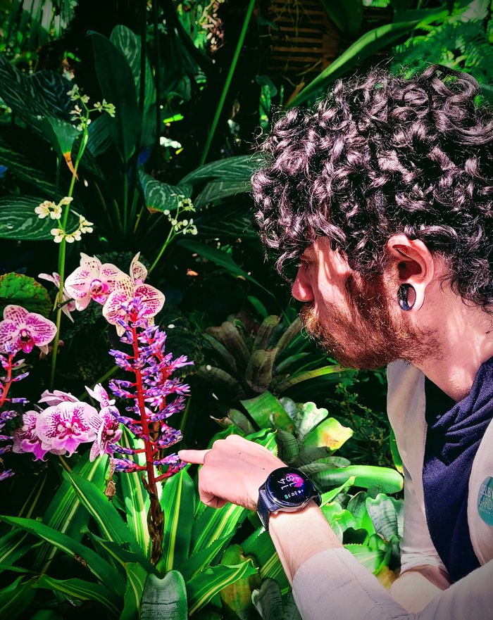 Zii in profile, pointing at a spike of pink and white orchid blooms in a lush greenhouse.